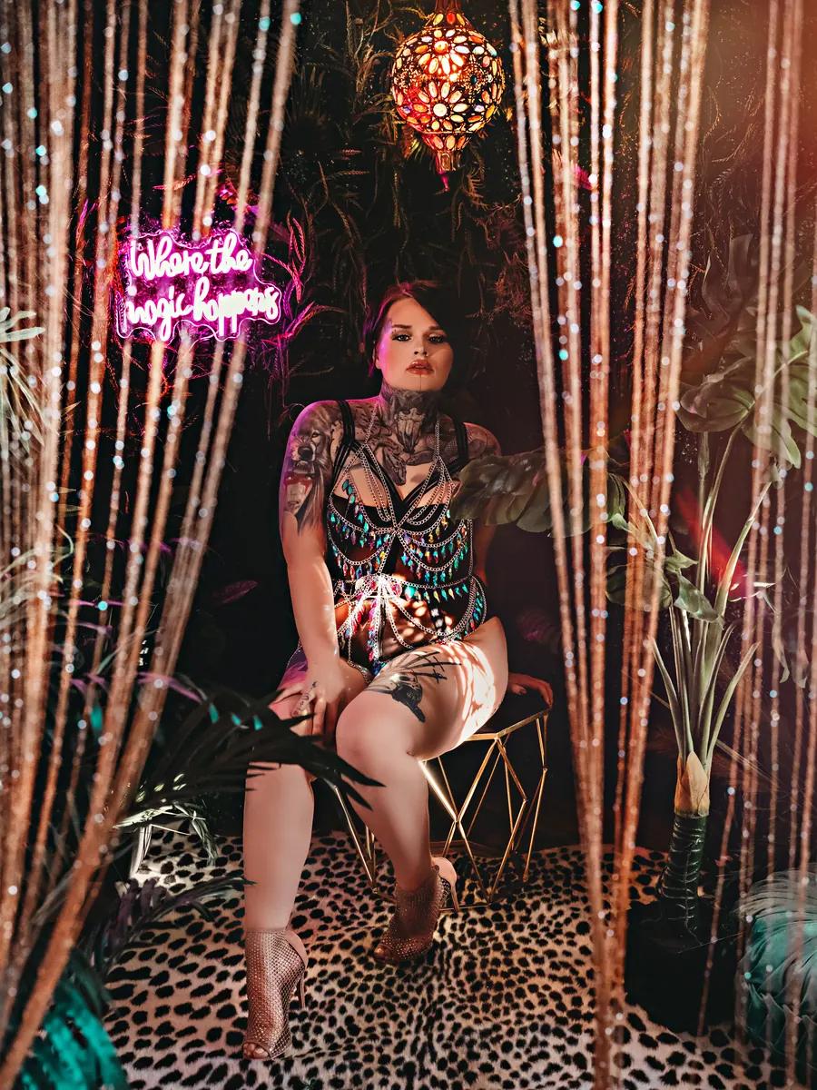 Tattooed client seated between beaded curtains on a leopard rug, under a pink neon sign
