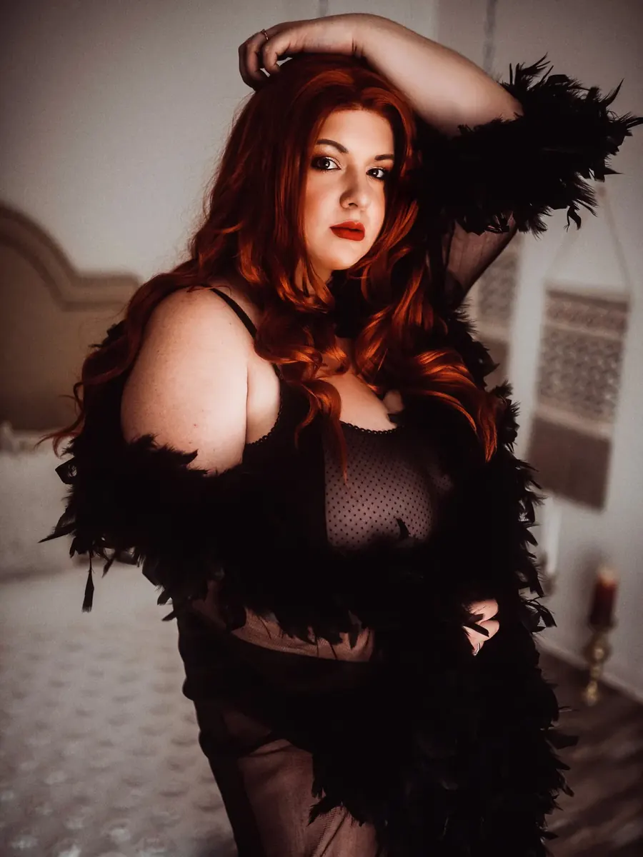 Client with long red hair in a sheer black feather-trimmed robe, one arm raised, in soft studio light