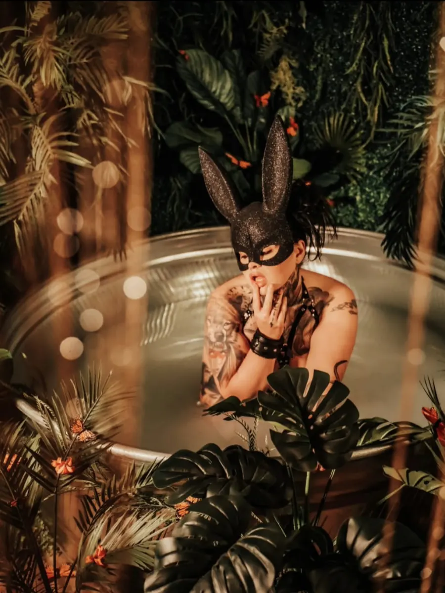 Tattooed client in a black bunny mask in a round tub surrounded by monstera leaves