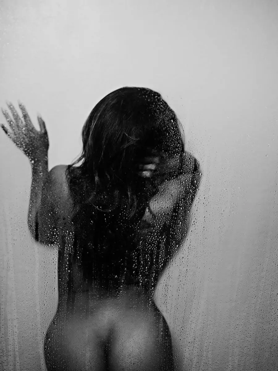 Black-and-white portrait of a client from behind, one hand on misted shower glass