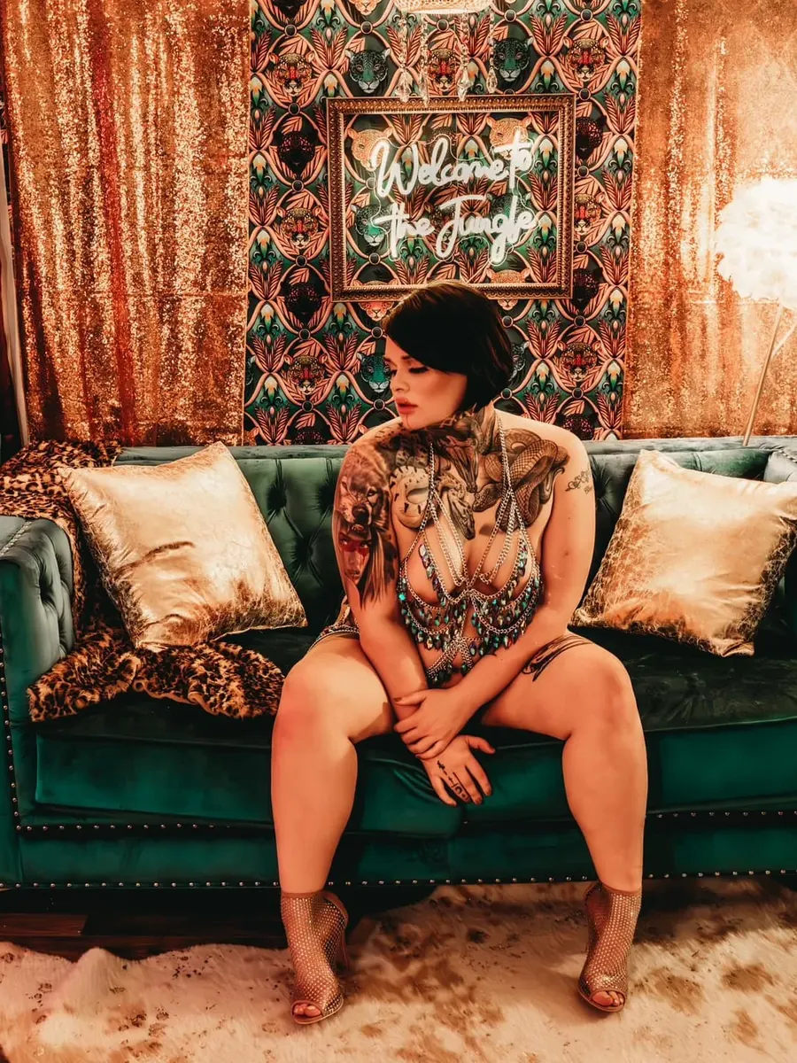 Tattooed client seated on an emerald velvet sofa against gold sequin drapes and patterned wallpaper