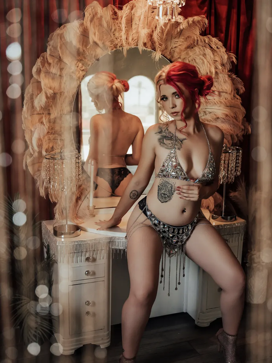 Red-haired client in a jewelled set at the feather-framed vanity, reflected in the mirror