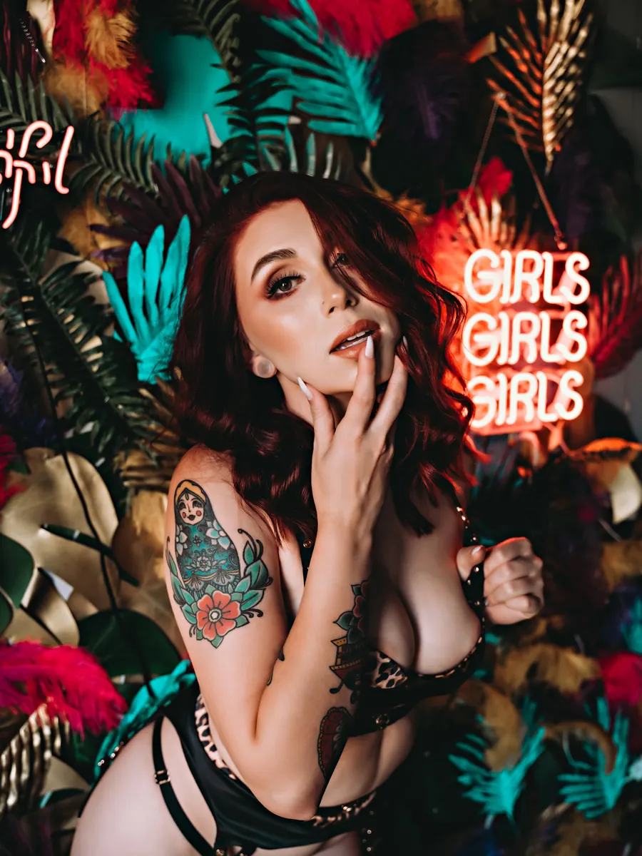 Tattooed client in black strappy lingerie against tropical floral wallpaper and a pink neon sign reading Girls Girls Girls