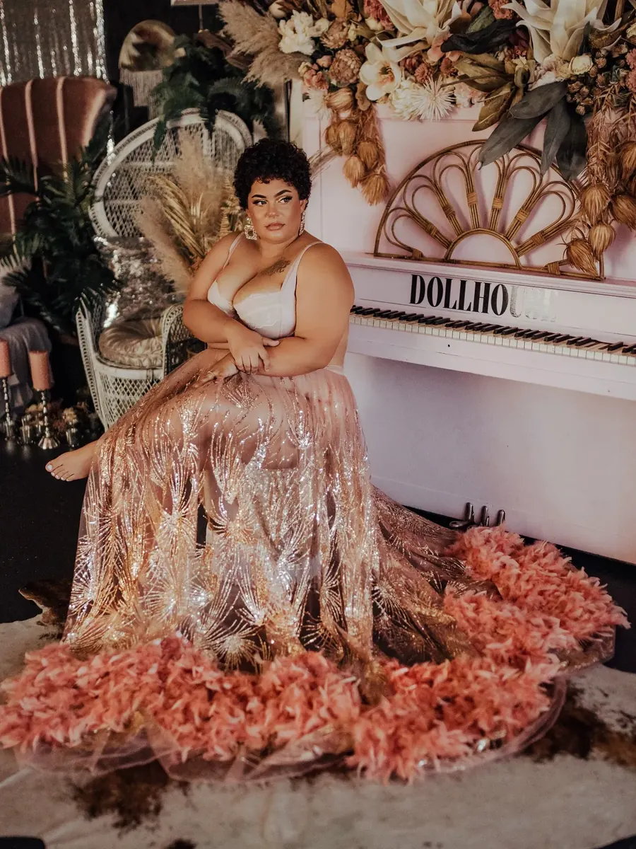 Client in a gold sequin gown with a pink feather hem, seated beside the white studio piano and a dried floral wall