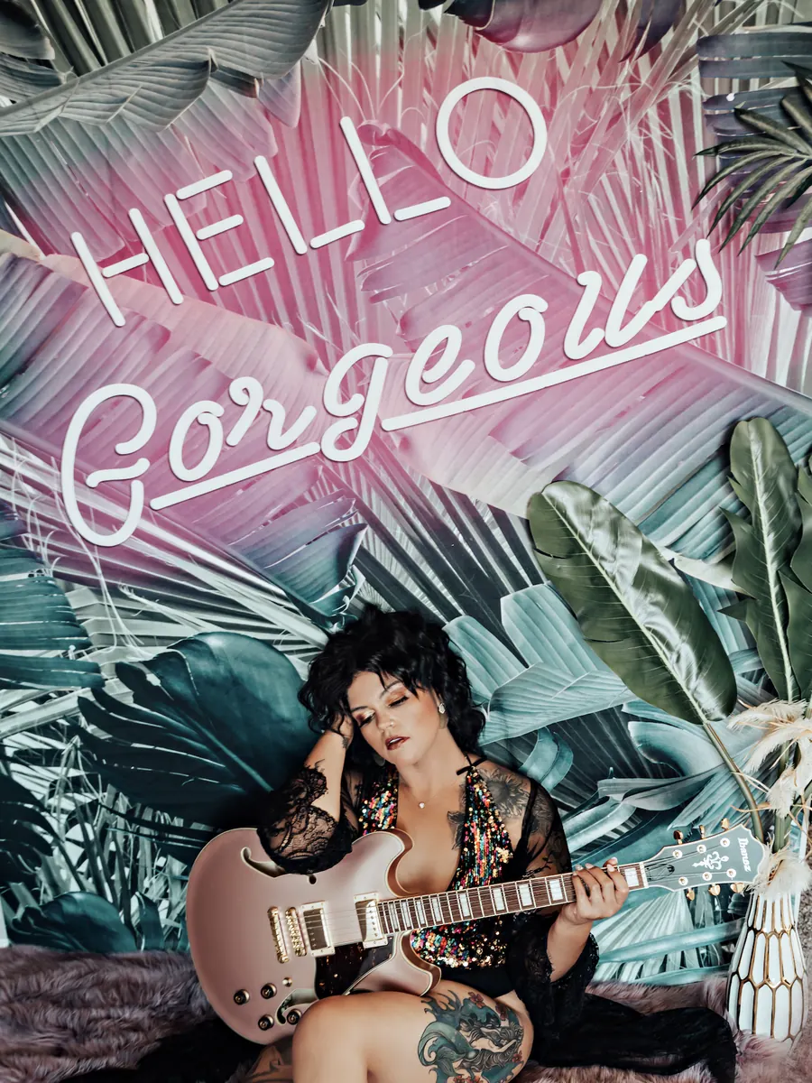 Client with a gold electric guitar beneath a pink neon Hello Gorgeous sign and tropical wallpaper