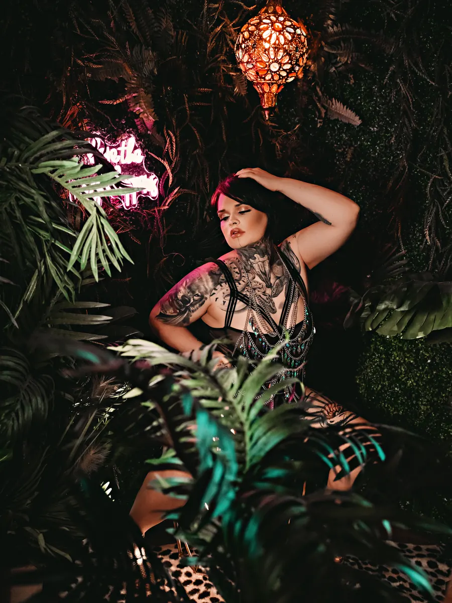 Client in a sheer lace robe among tropical plants beneath a lantern and pink neon