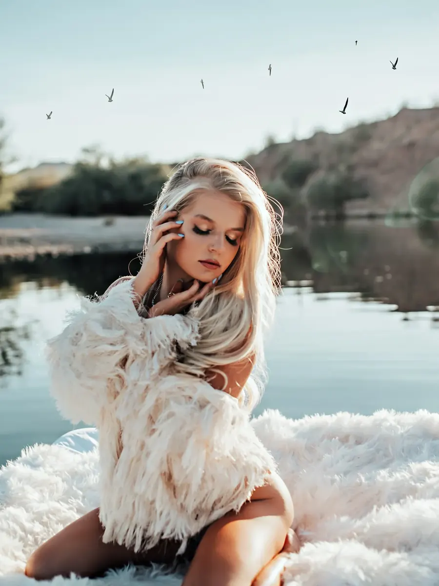 Blonde client kneeling on white fur at the edge of an Arizona lake, wrapped in a white feather jacket