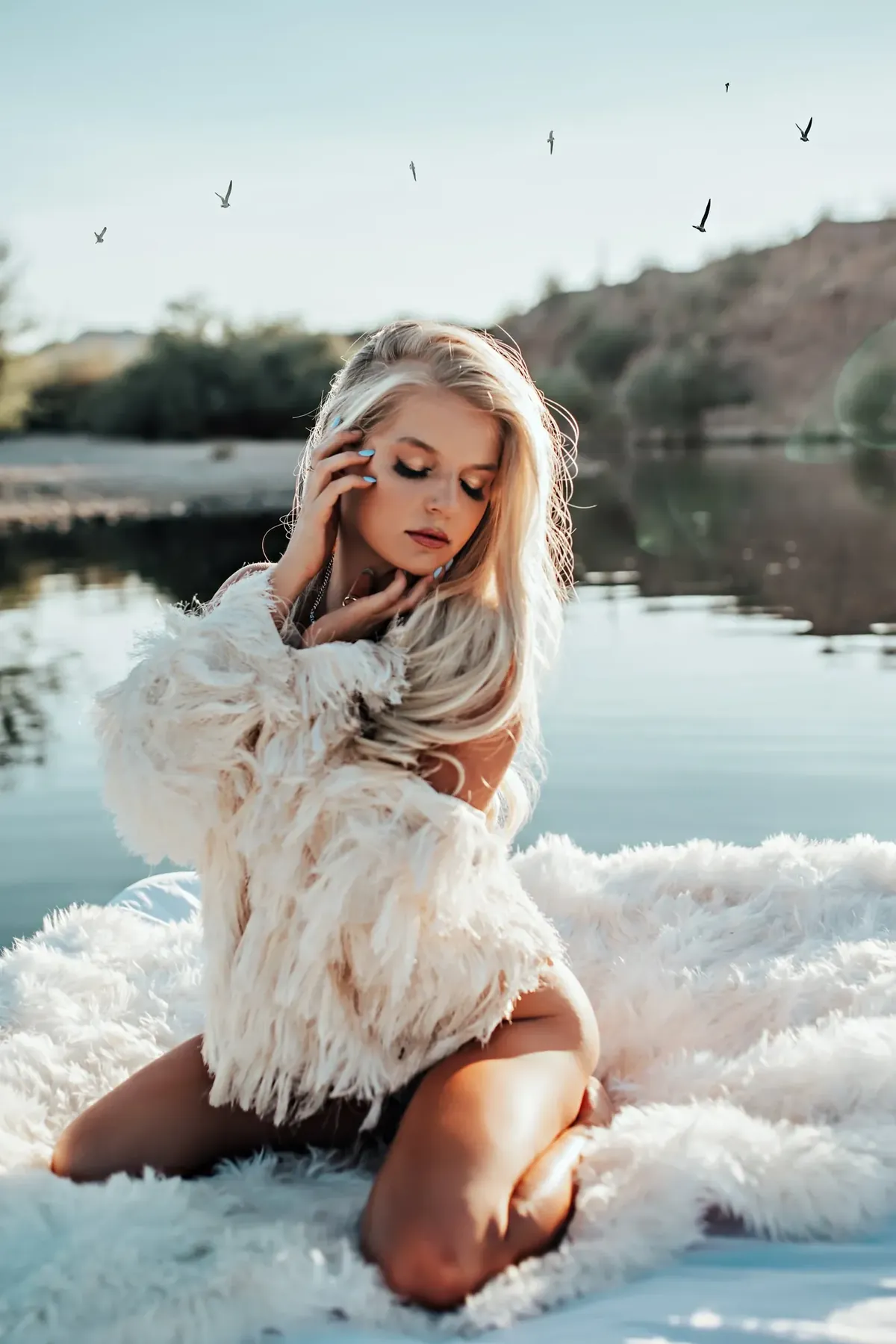 Client kneeling on white fur at the edge of an Arizona lake in a white feather jacket