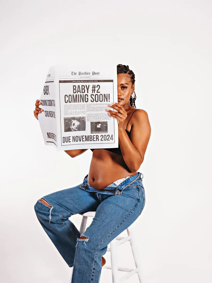 Expectant mother in denim seated on a white stool, holding a newspaper printed with a baby announcement