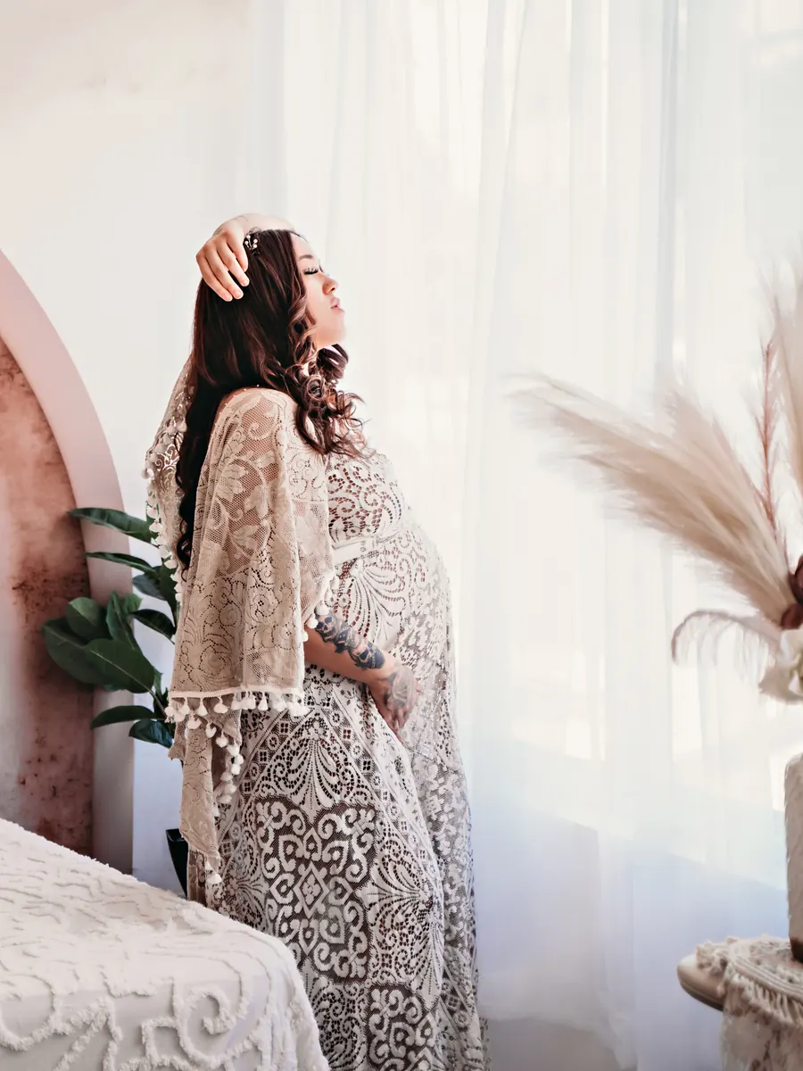 Expectant mother in a lace robe standing by the window beside a blush arched wall and bed