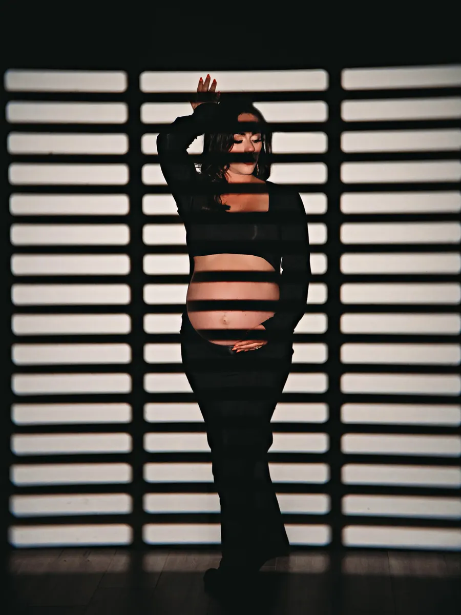 Silhouette of an expectant mother in a black gown against striped window-blind light