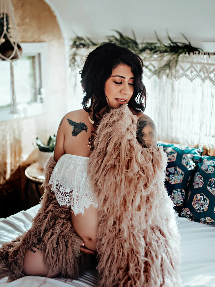 Expectant mother in a blush feather robe seated on a white bed