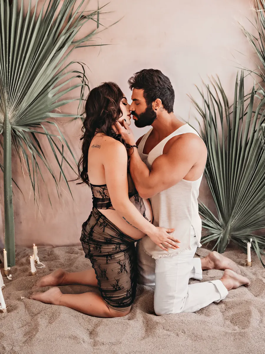 Expectant mother and her partner kneeling face to face on sand among palms and candles