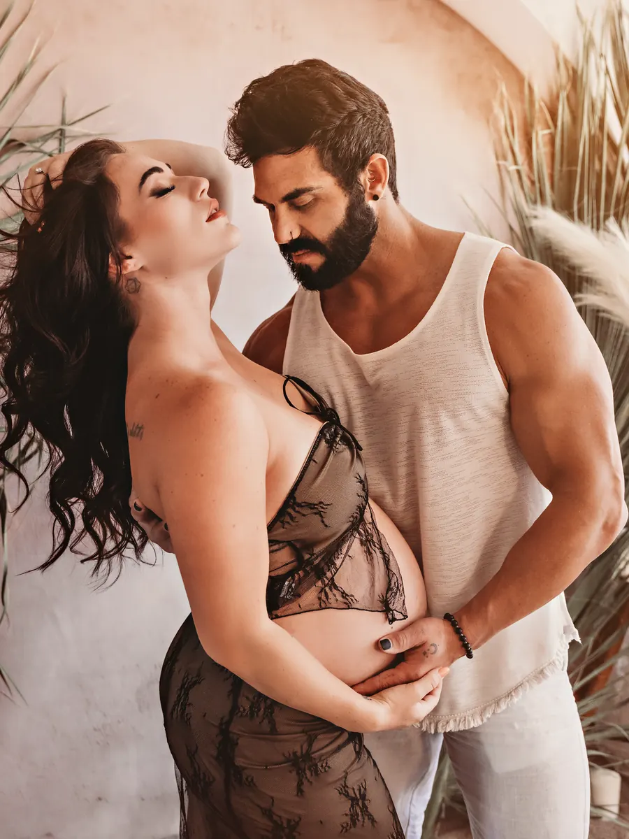Expectant mother in black lace with her partner’s hands resting on her belly, framed by dried palms