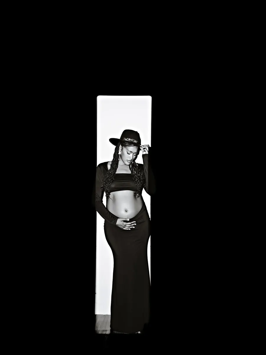 Expectant mother in a black hat and gown standing in a narrow doorway of light