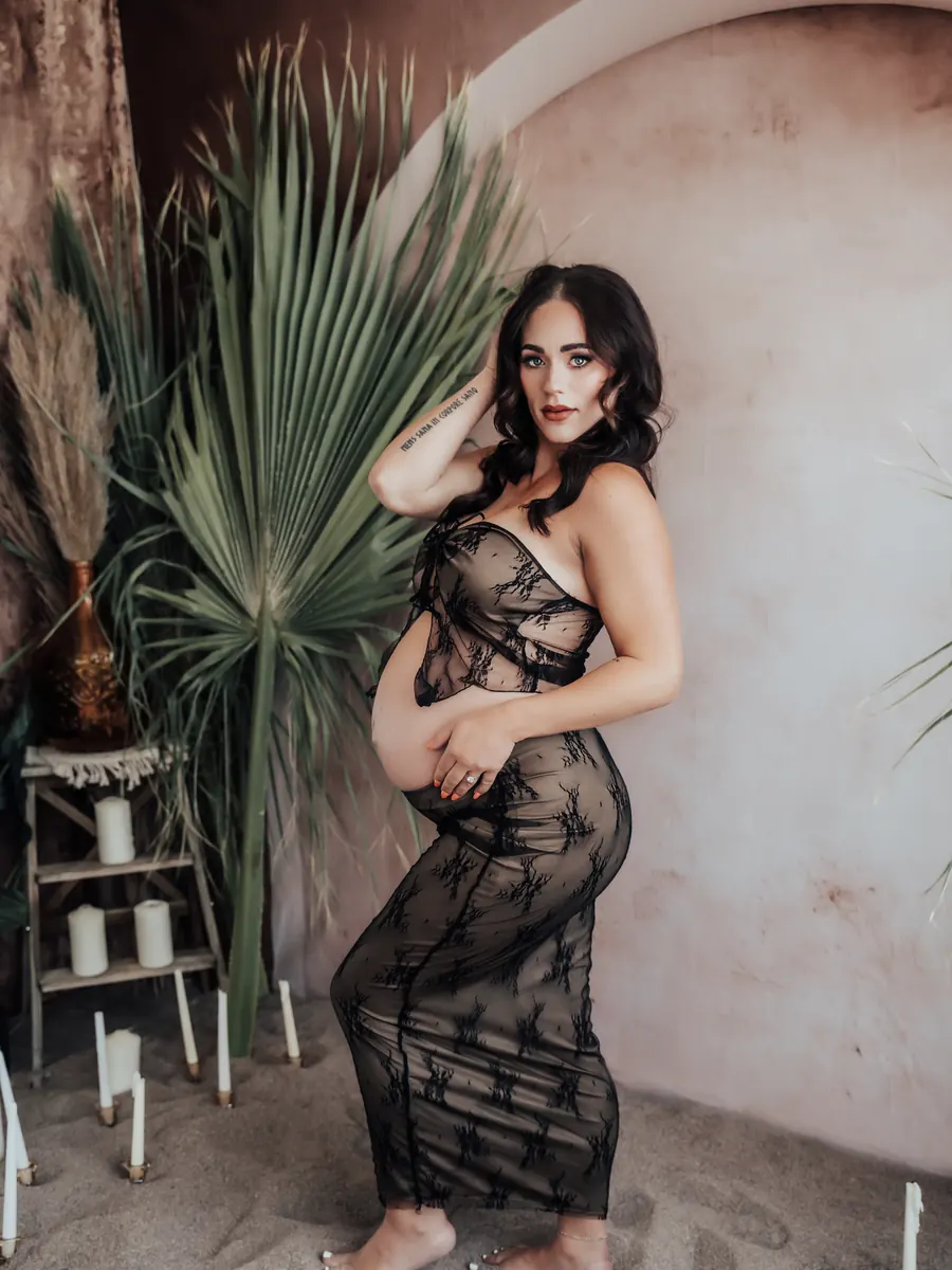 Expectant mother standing barefoot on sand in black lace among palms and candles
