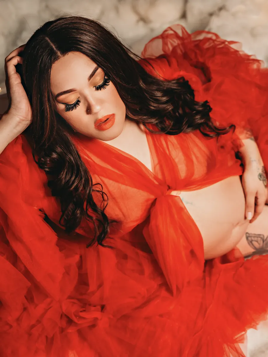 Expectant mother reclining in a red tulle robe on soft clouds