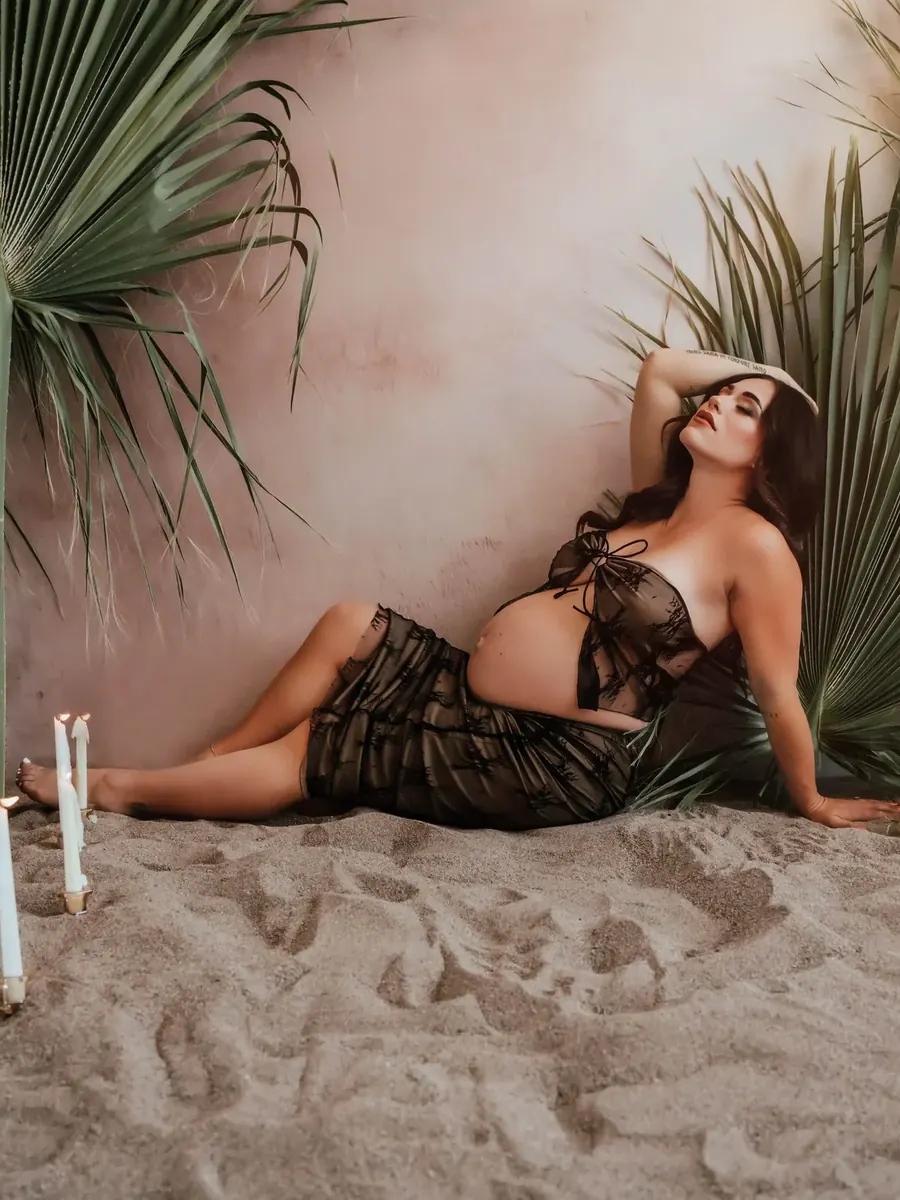 Expectant mother in black lace reclining on sand among palms and candles