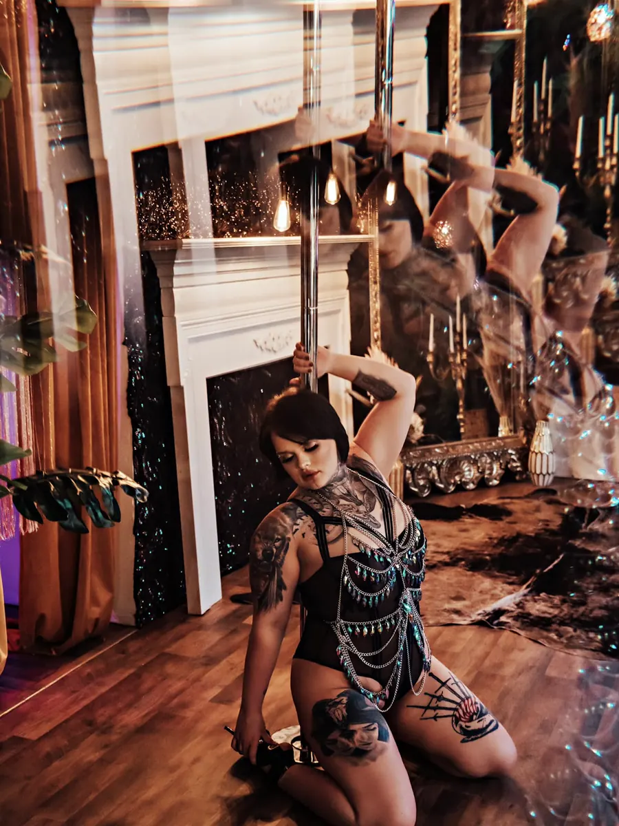 Tattooed client kneeling on a wood floor in a lace bodysuit, framed by mirrors and candlelight
