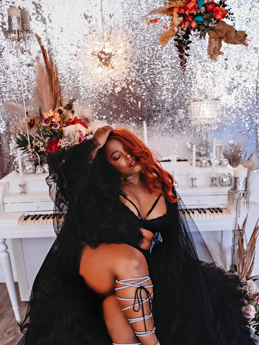 Red-haired client in a black feathered robe and lace-up heels beside the white studio piano