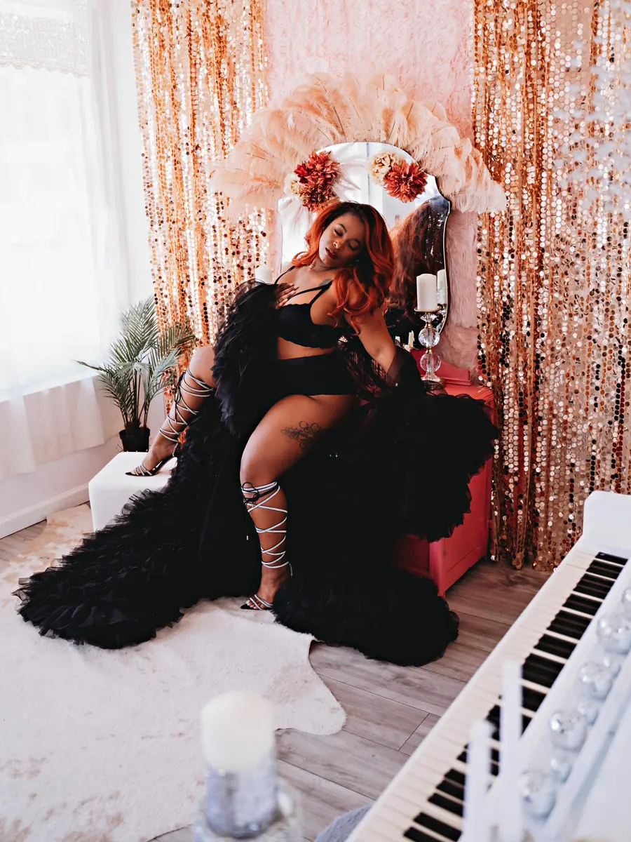 Client in a black feather robe reclining beside the white piano and gold sequin curtains