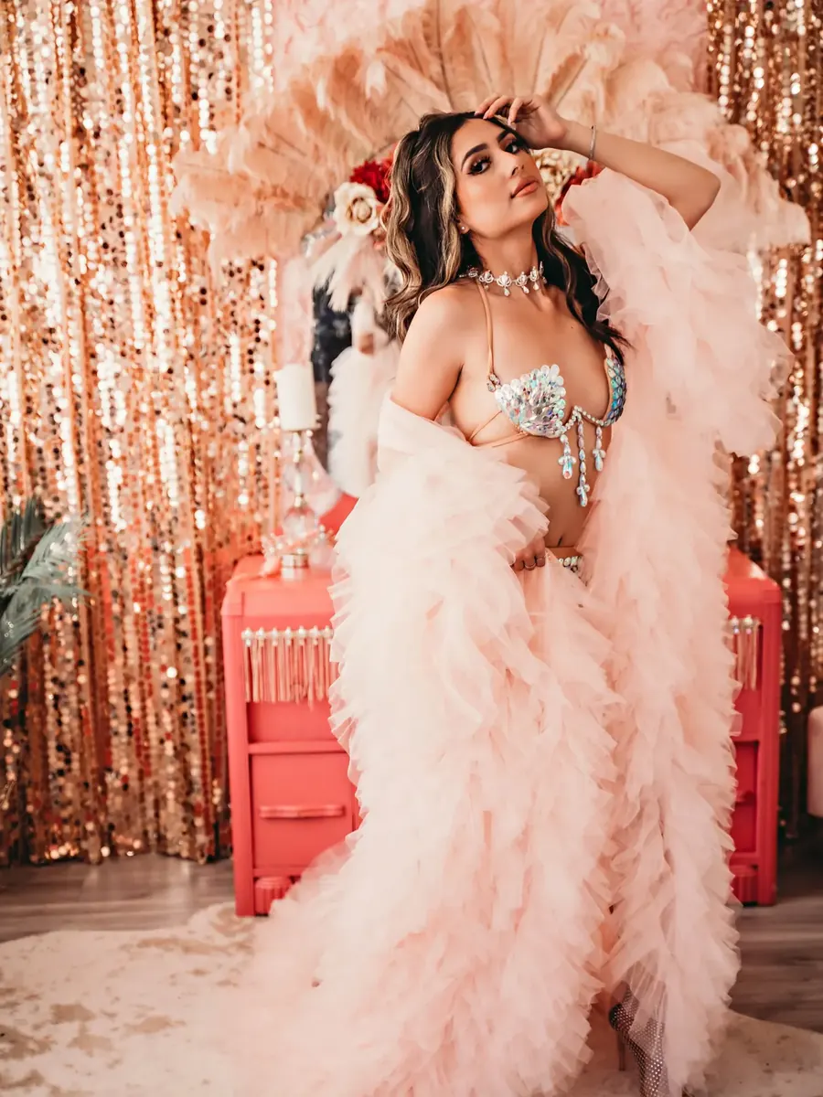 Client in a jewelled bralette and a floor-length blush tulle robe in front of a gold sequin wall