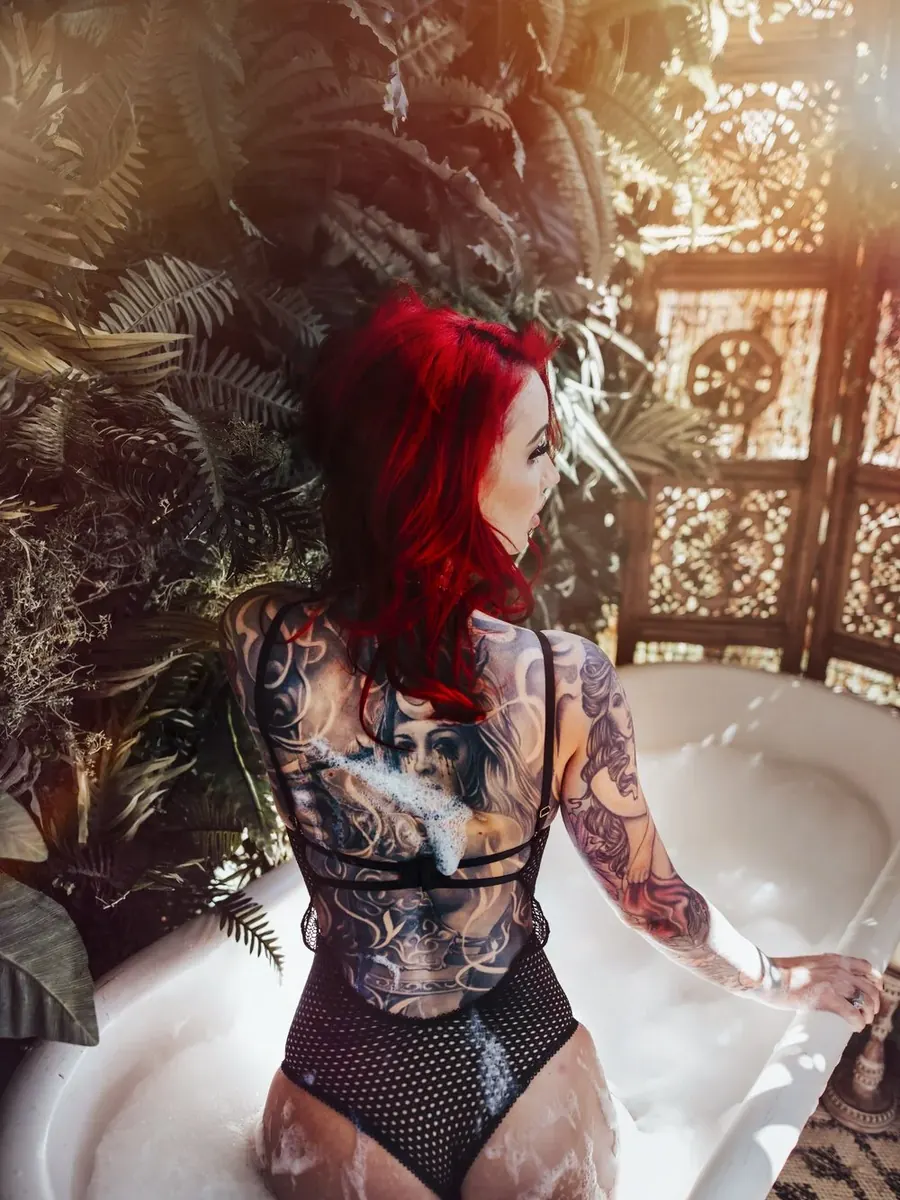 Red-haired client with a back full of tattoos kneeling in a milky bath among palms