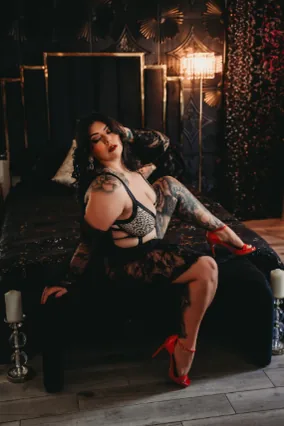 The same client lounging on the black velvet bed set in lace lingerie and red heels, under a gold chandelier sconce