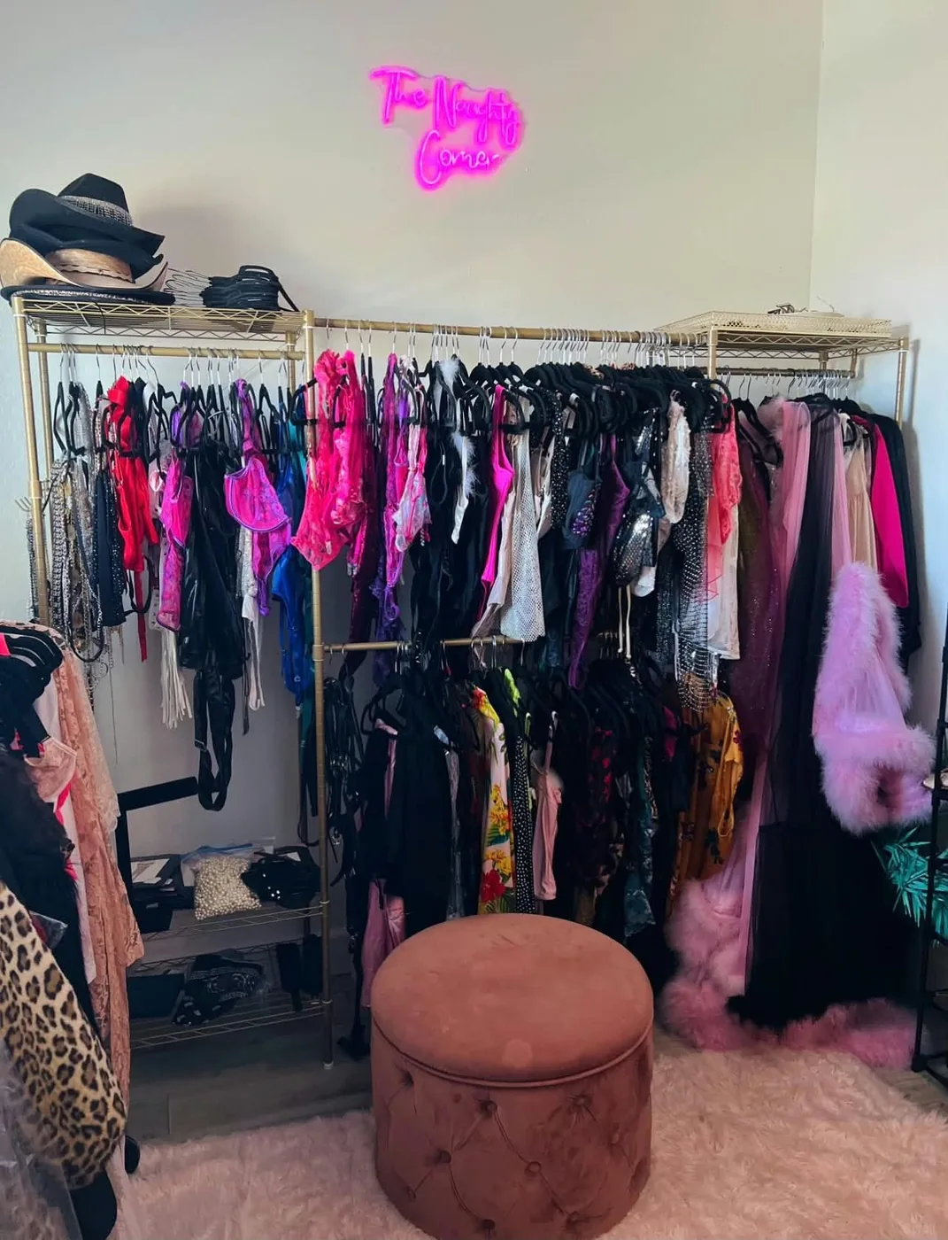 The client closet: gold racks of lingerie, robes and gowns beneath a pink neon sign, with a velvet ottoman and western hats