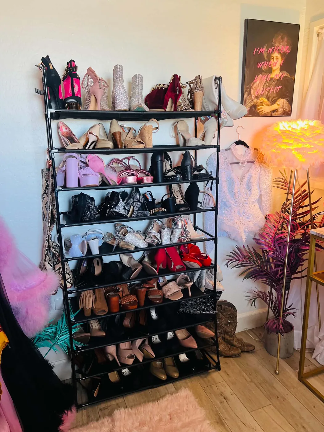 A tall rack of heels and boots in the client closet beside a feathered lamp and a framed portrait