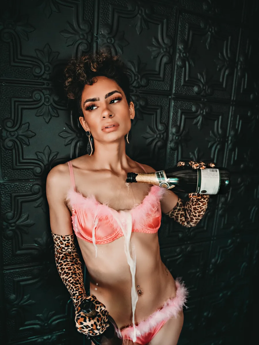 Client in pink feather lingerie and leopard gloves pouring champagne against a black tin wall